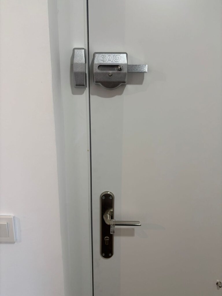 Puerta blanca con cerrojo de seguridad SAG plateado y maneta con bombín M&C instalados como doble sistema de cierre
